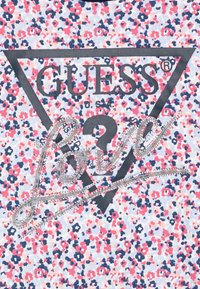 Biela košeľa s kvetinovým vzorom v ružovej, modrej a červenej farbe. Obsahuje veľký čierny trojuholník s nápisom "GUESS" a detailom z kryštálov "Love".