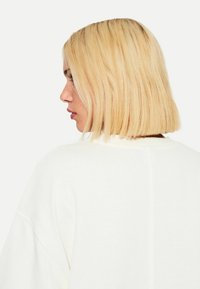 Felpa bianca con vestibilità rilassata, cuciture visibili e dettagli a coste, inquadrata di spalle con capelli biondi lisci e di lunghezza spalla.