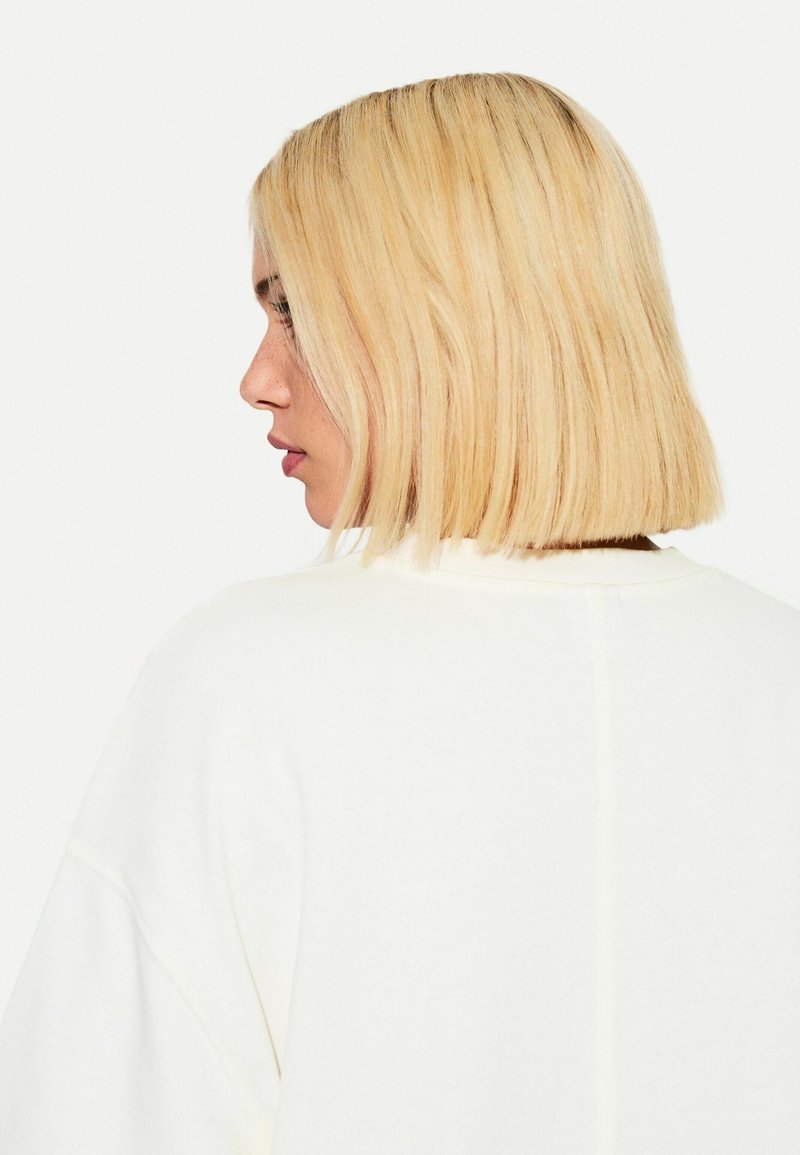 Felpa bianca con vestibilità rilassata, cuciture visibili e dettagli a coste, inquadrata di spalle con capelli biondi lisci e di lunghezza spalla.