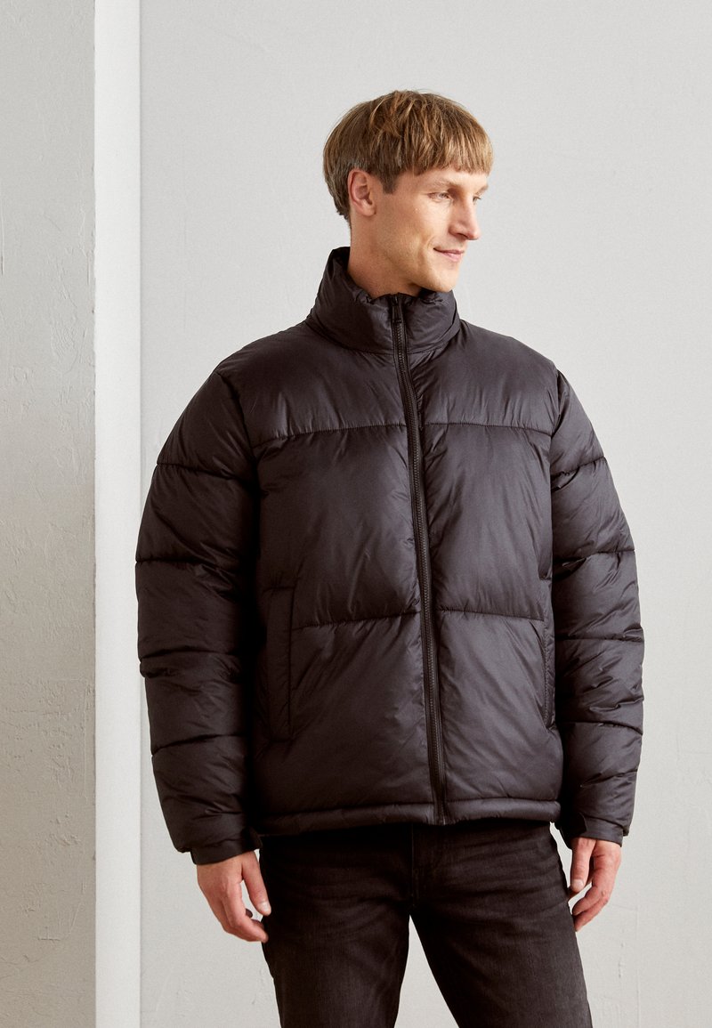 Veste doudoune noire avec un col haut, fermeture éclair et design matelassé. Fabriquée en matériau léger et isolant avec des poches avant.