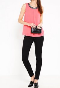 Blouse sans manches rose avec un col en perles, associée à un pantalon skinny noir et des chaussures plates noires. Pochette noire tenue dans une main.