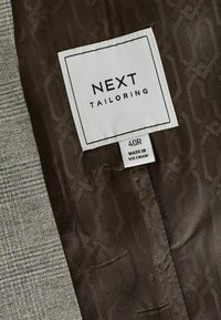 Interior de chaqueta a medida con un patrón de houndstooth gris claro y un forro de satén marrón oscuro con un logo grabado y etiqueta de cuidado.