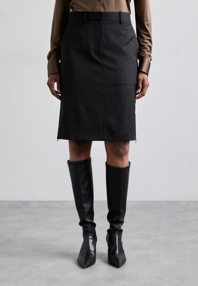 Jupe crayon noire à texture lisse, avec un zip frontal et une silhouette ajustée, associée à de grandes bottes en cuir noires à bout pointu.