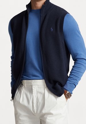 Uomo che indossa un gilet blu navy con cerniera sopra una camicia a maniche lunghe blu, pantaloni color crema e un orologio da polso in pelle marrone.