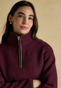 Bordo vilnas džemperis ar augstu apkakli, pus-zips ar zelta detaļām un kontrastējošām brūnām akcentiem pie aprocēm un apkakles. Mazs logo uz krūtīm.