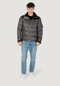 Giovane uomo in giacca imbottita grigia con accenti marroni, jeans azzurri chiari e sneakers bianche, in piedi davanti a uno sfondo semplice e chiaro.