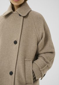 Taupe wollen jas met een grote kraag, zwarte knopen en zijzakken. Heeft een ontspannen pasvorm en een gestructureerde stof.