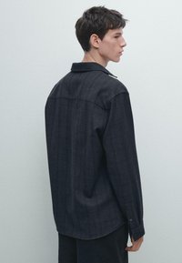 Unge mand med kort, mørkt hår iført en løs, mørkegrå langærmet skjorte, set fra ryggen og siden mod en ensfarvet lys baggrund.