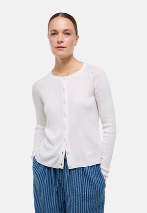 Femme à la peau claire et aux cheveux bruns porte un cardigan blanc boutonné et un pantalon bleu rayé, debout les mains dans les poches devant un fond uni.