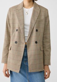 Blazer beige con motivo a quadri marrone e rosso, design doppiopetto, bottoni neri e taschino sul petto; indossato sopra una camicia bianca.