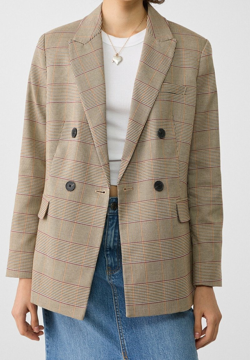 Blazer beige con motivo a quadri marrone e rosso, design doppiopetto, bottoni neri e taschino sul petto; indossato sopra una camicia bianca.