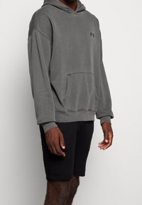 Harmaa hupullinen collegepaita kengurun taskulla ja resorireunuksilla hihoissa. Yhdistetty mustiin shortseihin, joissa on rento istuvuus ja pehmeä tekstuuri.