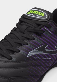 Μαύρο και μωβ αθλητικό παπούτσι Joma Maxima με μαύρα κορδόνια, λογότυπο και κείμενο που αναγράφει «Άριστη Εφαρμογή» και «Σύστημα Εξαερισμού» στο πλάι.
