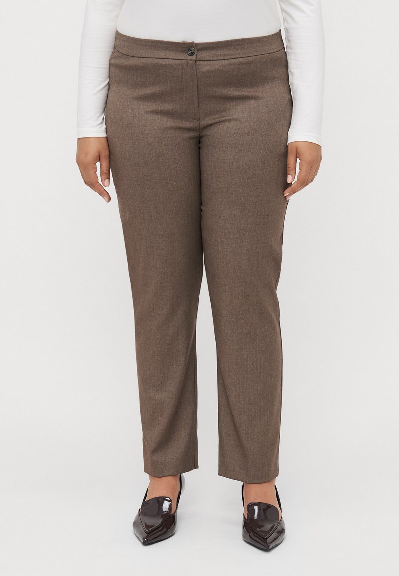Pantalons marron ajustés avec une texture lisse, coupe slim et fermeture à un seul bouton. Portés avec des chaussures sombres à bout pointu et brillantes.