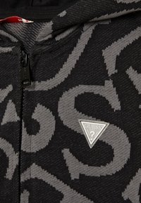 Felpa con cappuccio nera con un audace motivo di logo grigio. Un accento triangolare argentato con il logo "GUESS" è posizionato sul lato sinistro, vicino alla cerniera.