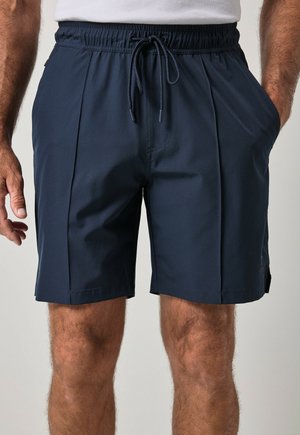 Man draagt donkerblauwe shorts met trekkoord en zijzakken en een wit overhemd, handen ontspannen langs het lichaam.