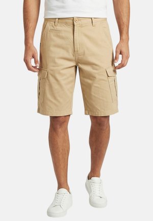 Homme portant un short cargo beige et des baskets blanches, debout, les mains détendues le long du corps, sur un fond uni.