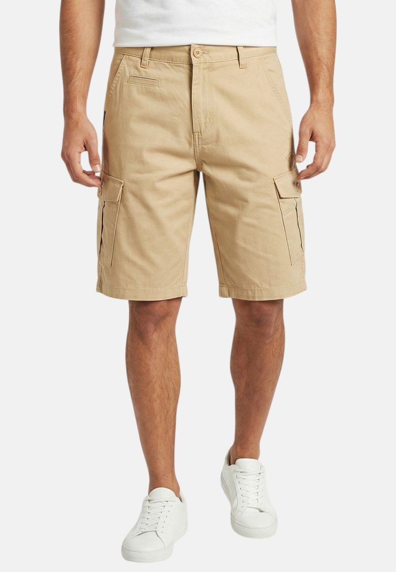 Homme portant un short cargo beige et des baskets blanches, debout, les mains détendues le long du corps, sur un fond uni.