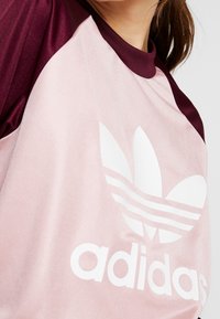 Rosa och mörkröd sporttröja med korta ärmar, med en stor vit Adidas-logotyp och den ikoniska tre-stripesdesignen. Slät tygstruktur.