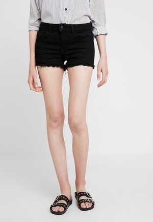 Persona con shorts vaqueros negros de dobladillo deshilachado, camisa a rayas con botones y mangas remangadas, y sandalias negras con hebilla, de pie contra un fondo gris.
