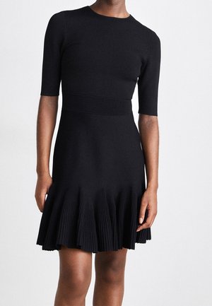 Femme portant une robe noire ajustée avec des manches mi-longues jusqu'aux coudes et un ourlet de jupe plissé, debout devant un fond clair uni.
