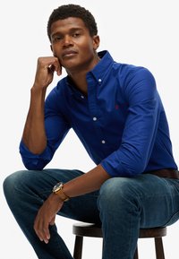 Een blauwe button-up shirt met opgerolde mouwen, voorzien van een klein rood logo. Draagt het met donkere denim jeans en een bruine leren horloge, zittend op een kruk.