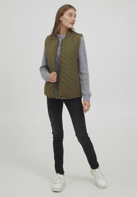 Olivgrön kviltad väst med dragkedja fram, hög krage och sidofickor, kombinerad med en grå stickad tröja och svarta slim jeans. Vita sneakers.