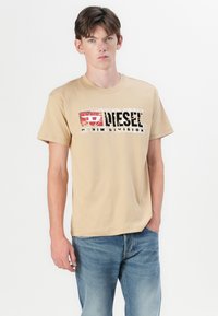 Smėlinis medvilnės marškinėliai su nusidėvėjusiu juodu ir raudonu "Diesel Denim Division" piešiniu priekyje. Trumpi rankovės ir klasikinis apykaklės dizainas.