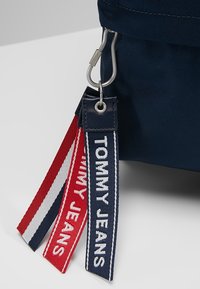 Sac en tissu bleu marine avec garnitures en argent, doté de trois étiquettes en tissu rouges, blanches et bleu marine, marquées "TOMMY JEANS" et de rayures décoratives.