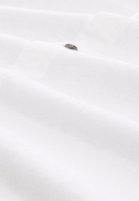 Weißer Stoff mit glatter Textur, der zarte Falten und ein auffälliges metallisches Knopfdetail in der Mitte aufweist.