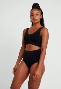 Punutud juustega naine, kes kannab musta spordirinnahoidjat ja kõrge vöökohaga aluspükse, seisab ühetaolise heleda halli taustaga.