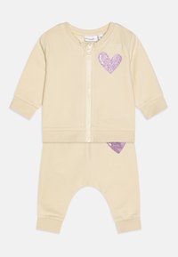 Beige cotton zip-up jacket and pants set with ribbed cuffs. Features a purple glitter heart on the jacket and pant. Soft texture.