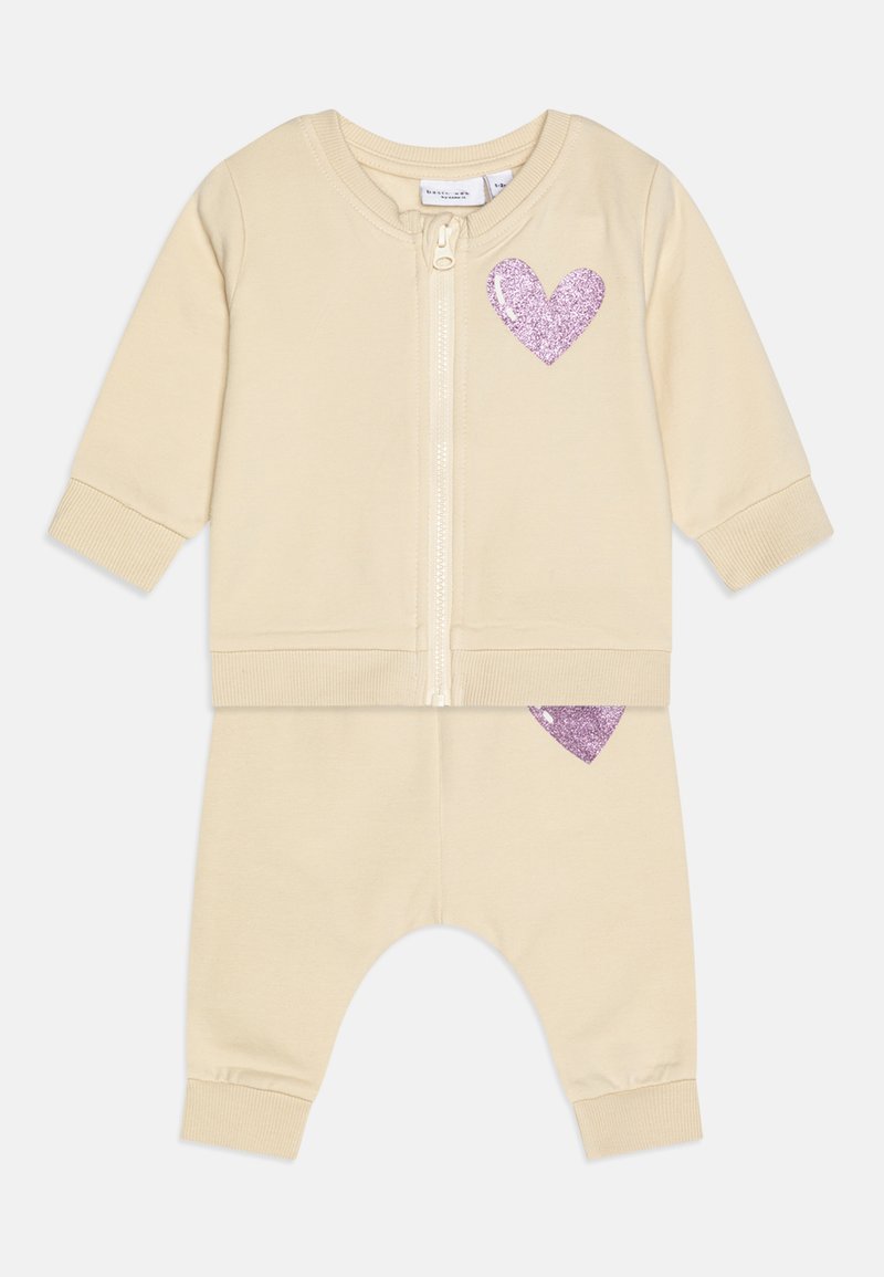 Beige cotton zip-up jacket and pants set with ribbed cuffs. Features a purple glitter heart on the jacket and pant. Soft texture.