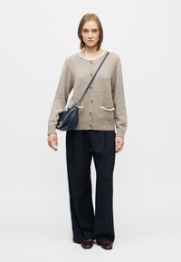 Beige knitted cardigan with a scalloped neckline, front button closure, and two pockets. Paired with navy wide-leg pants and a dark handbag.