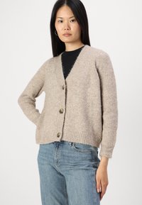 Beige gebreide cardigan met een v-hals, lange mouwen en knoopsluiting. Zachte textuur en ribgebreide zoom. Gemaakt over een zwarte top en blauwe jeans.