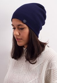 Navy ribbed beanie with a soft texture and a logo patch on the side, worn with a light-colored knitted sweater.