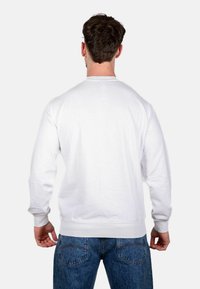 Felpa bianca con colletto rotondo, maniche lunghe, in cotone, con polsini e orlo a costine, indossata con jeans in denim blu vista da dietro.