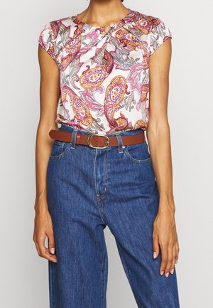 Blusa de manga corta con un patrón de paisley en rosa, amarillo y azul sobre un fondo blanco; combinada con pantalones de mezclilla azules de cintura alta y un cinturón marrón.