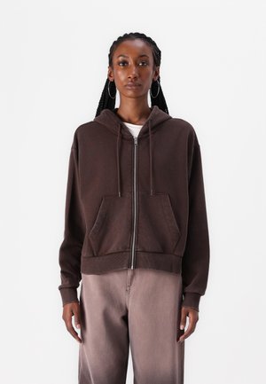 Felpa marrone con zip, cappuccio regolabile con cordino, tasche frontali e polsini a costine. Indossata sopra una maglietta bianca, abbinata a pantaloni marrone chiaro.