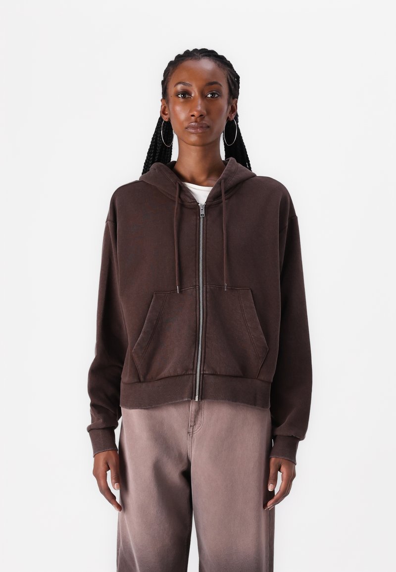 Felpa marrone con zip, cappuccio regolabile con cordino, tasche frontali e polsini a costine. Indossata sopra una maglietta bianca, abbinata a pantaloni marrone chiaro.