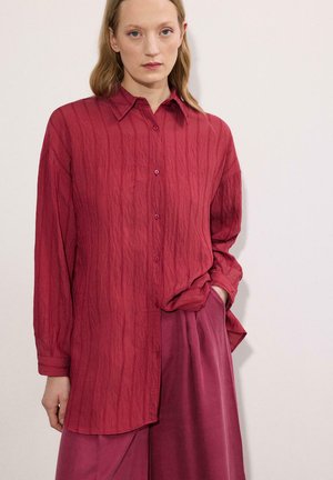 Γυναίκα που φοράει άνετο κόκκινο μακρυμάνικο πουκάμισο με υφή και παντελόνια με φαρδιά μπατζάκια σε ίδιο χρώμα, στέκεται μπροστά σε ένα απλό ανοιχτό φόντο.