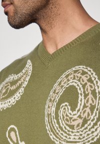 Alyvuogių žali mezginys su V formos kaklu, puoštas sudėtingais baltais paislio raštais ir lapų motyvais, su ribotu apykaklės ir apačios dizainu.