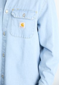 Gai zils džinsu krekls ar divām krūšu kabatām, sudraba pogām un dzeltenu un balta Carhartt logo etiķeti uz kreisās kabatas.