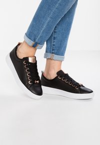 Černé tenisky s hladkým koženým povrchem, očky v růžovém zlatě a bílou gumovou podrážkou. Obsahují texturované akcenty poblíž paty.