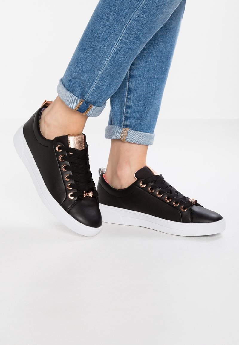 Černé tenisky s hladkým koženým povrchem, očky v růžovém zlatě a bílou gumovou podrážkou. Obsahují texturované akcenty poblíž paty.