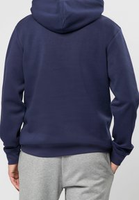 Personne portant un sweat à capuche bleu marine et un pantalon de jogging gris clair, vue de dos, debout avec les mains détendues le long du corps.