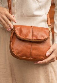 Hnědá kožená crossbody taška s uzávěrem na zip, zaobleným tvarem a hladkým povrchem s klopou. Nastavitelný popruh se zlatými doplňky.