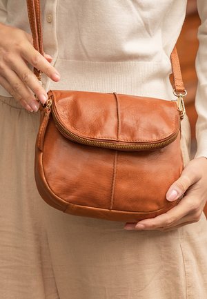 Torebka crossbody w kolorze brązowym z zamkiem błyskawicznym, okrągłym kształtem i gładką teksturą z klapą. Regulowany pasek z złotymi akcentami.