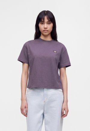 Jeune femme aux cheveux raides et foncés, portant un t-shirt violet à manches courtes et un jean bleu clair, debout devant un fond uni.