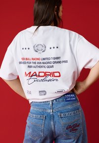 Woman wearing white Red Bull Racing Madrid Grand Prix limited edition T-shirt and blue jeans with Red Bull Racing branding, standing against red background.